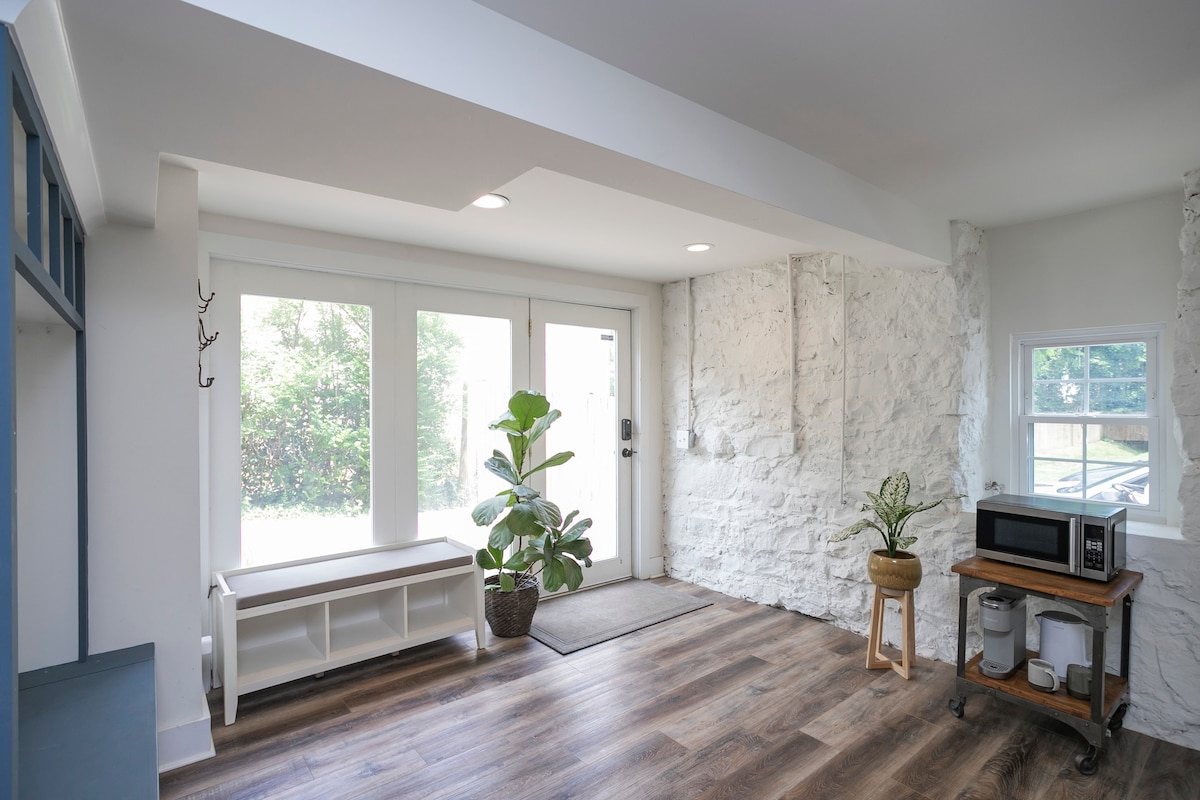 An airy mudroom features stone accent walls and large windows allowing natural light. A potted plant is situated by the entrance, with a small wooden table holding a microwave and additional storage on the opposite wall. The flooring is a warm wood tone.