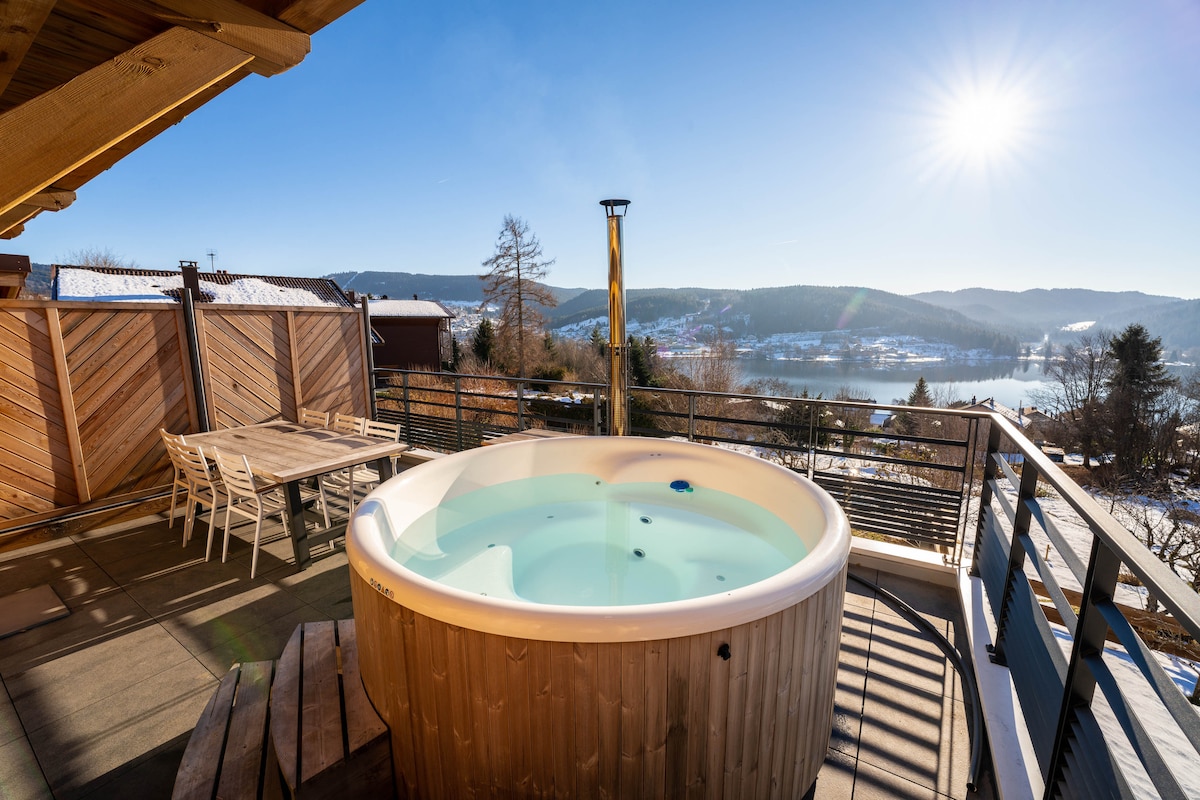 A private hot tub is positioned on a sunlit terrace, offering scenic views of the lake and surrounding landscape. A large wooden table is nearby, surrounded by several chairs, creating a space for outdoor dining or relaxation.