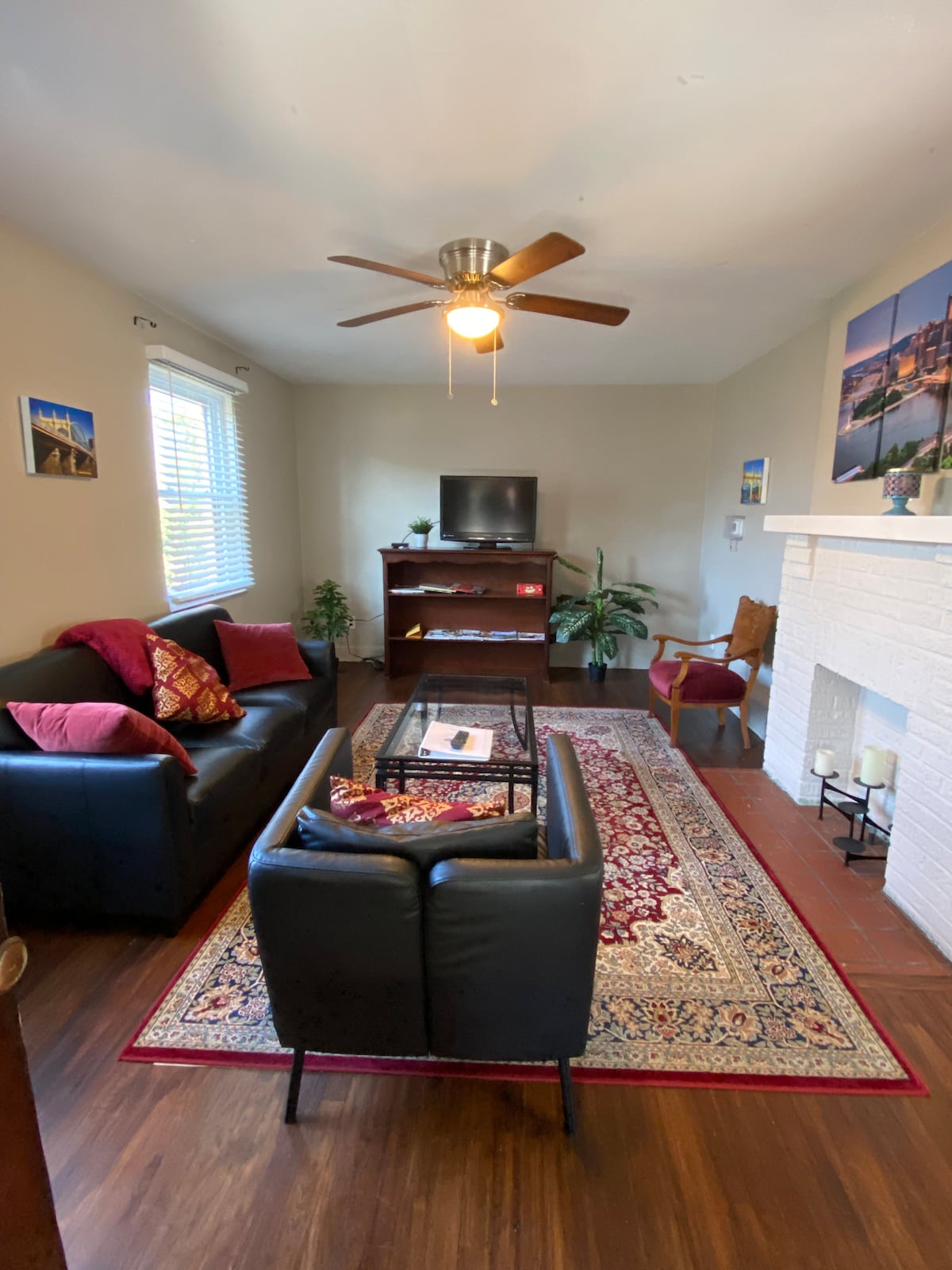 The living room features a comfortable arrangement of dark furniture, including a sofa and chairs, complemented by decorative pillows. A coffee table sits on a patterned area rug. Natural light enters through windows adorned with blinds, and a television is positioned against a white brick wall.