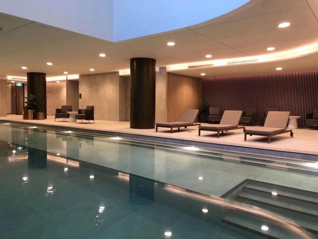 An indoor swimming pool is surrounded by comfortable lounge chairs. The sleek design features contemporary finishes, with soft lighting enhancing the tranquil atmosphere. Planted arrangements and decorative walls add to the elegance of the space, while the water's surface reflects the ambient light.