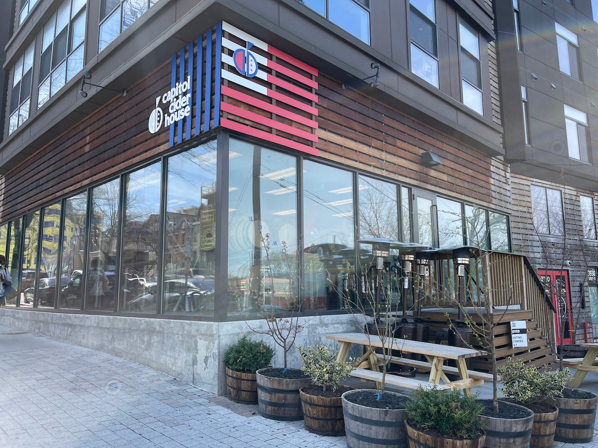 The exterior of a contemporary building is visible, featuring large windows and a distinctive signage with red, white, and blue stripes. Outdoor seating is provided with wooden benches surrounded by landscaped planters containing small shrubs.