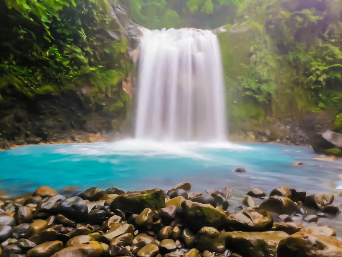 A serene waterfall cascades over rocks into a vibrant turquoise pool, surrounded by lush greenery. Smooth stones line the foreground, creating a natural border. The mist from the falling water adds a sense of tranquility to the scene.
