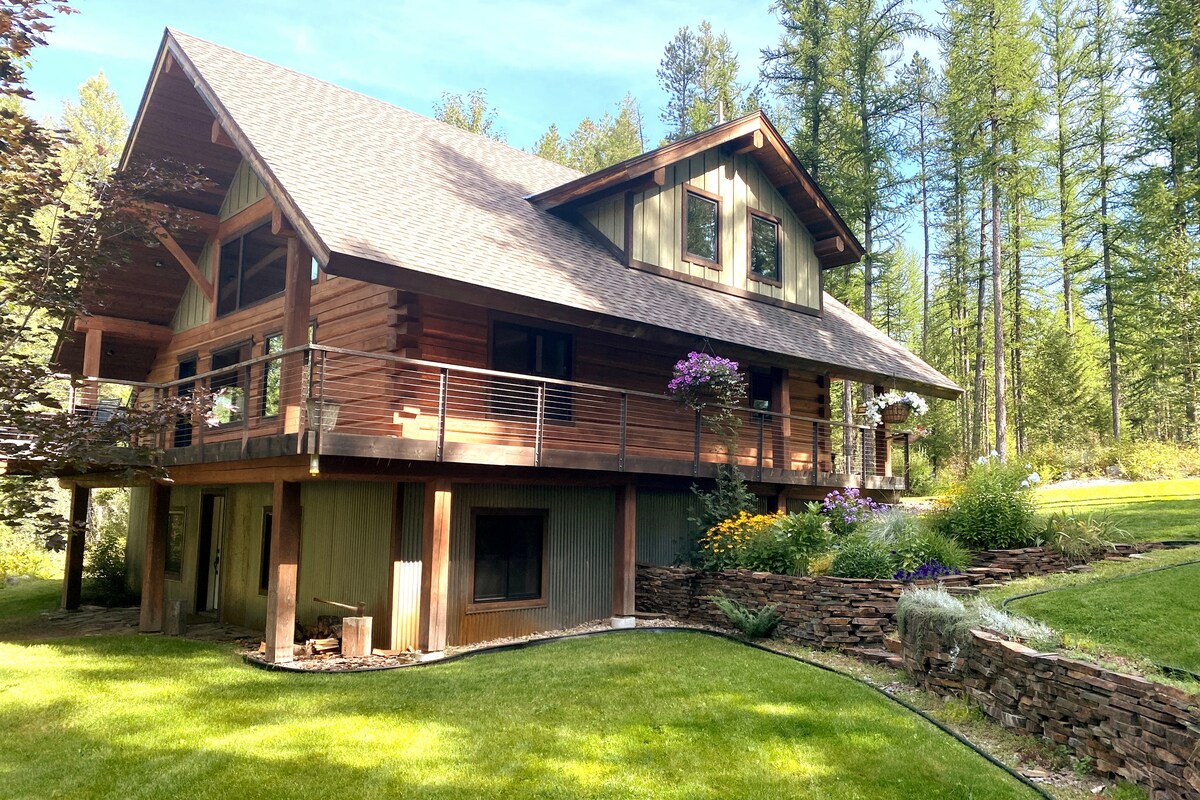 The log home features a multi-level design, surrounded by lush greenery and wildflowers. A spacious deck extends from the front, complemented by a railing and potted plants, offering a scenic view of the landscaped yard and tall trees in the background.