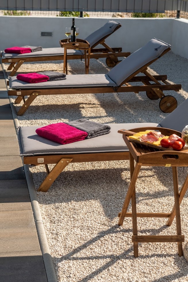Four lounge chairs with soft grey cushions are positioned on a gravel area, each adorned with a plush red and grey towel. A wooden table set beside one chair holds a tray featuring fresh apples and a snack, creating a relaxing outdoor space.