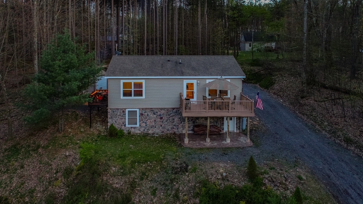 The exterior of the home is seen from a distance, surrounded by trees in a natural setting. A spacious balcony is visible, adorned with outdoor seating and an American flag. A gravel driveway leads up to the building, which features a mix of stone and siding.