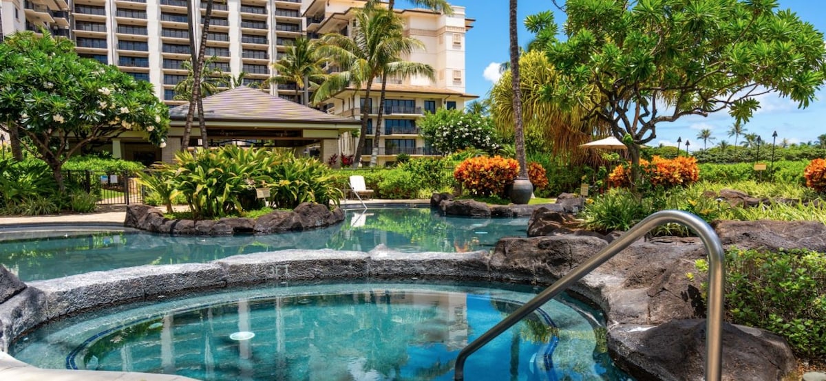 A tranquil spa area features a hot tub surrounded by lush tropical landscaping and colorful flowers. Nearby, a pathway leads to a larger pool area, with palm trees and vibrant greenery creating a serene atmosphere.