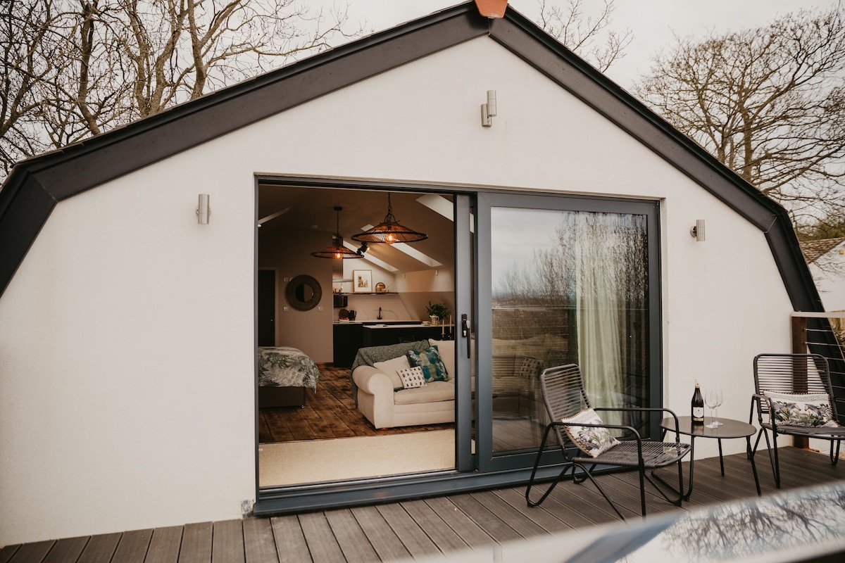 An open-plan loft apartment is visible through large sliding glass doors, inviting natural light. A comfortable sofa and kitchen area can be seen. A deck features two outdoor chairs, reflecting a serene outdoor space for relaxation.