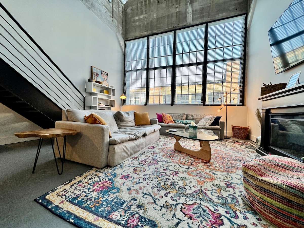 The spacious living area features large windows that allow ample natural light. A comfortable couch, styled with decorative pillows, and a unique coffee table are positioned on an intricately patterned rug. A small side table and a cozy pouf add to the inviting atmosphere.