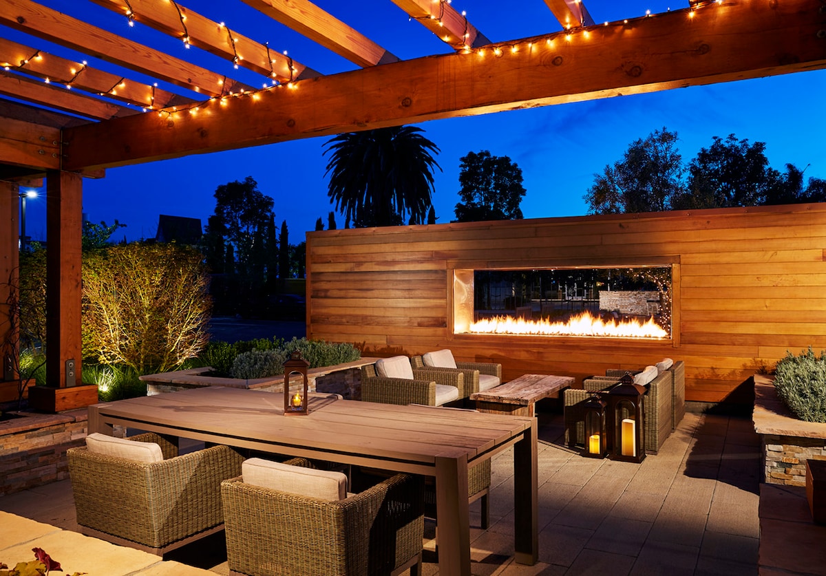 An outdoor lounge area is illuminated by string lights under a wooden pergola. A long dining table is surrounded by woven chairs, while a modern fireplace is integrated into a wooden wall, creating a calming ambiance. Lush greenery frames the space.