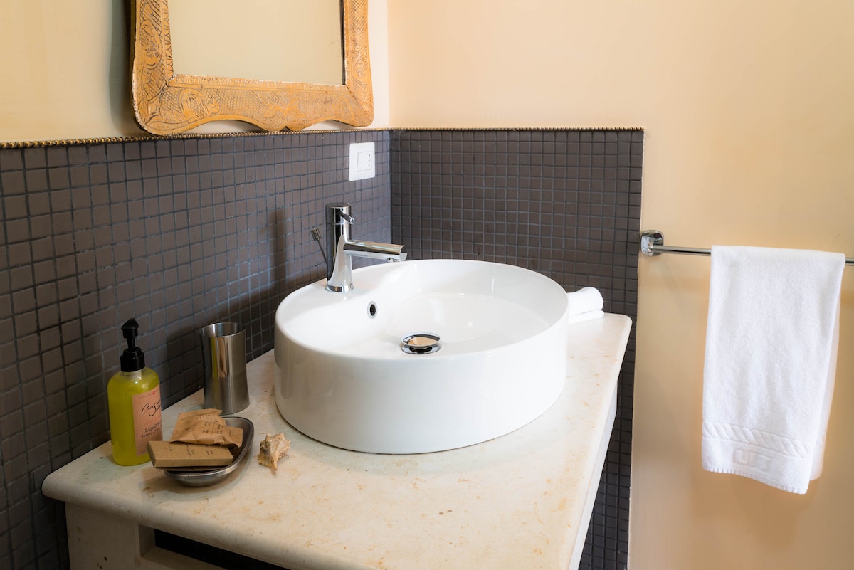 A modern bathroom sink is centered on a textured countertop, featuring a round basin and a sleek faucet. A wall-mounted towel bar holds a neatly hung white towel. Decorative items, including soap and toiletries, are arranged on the surface beside the sink.