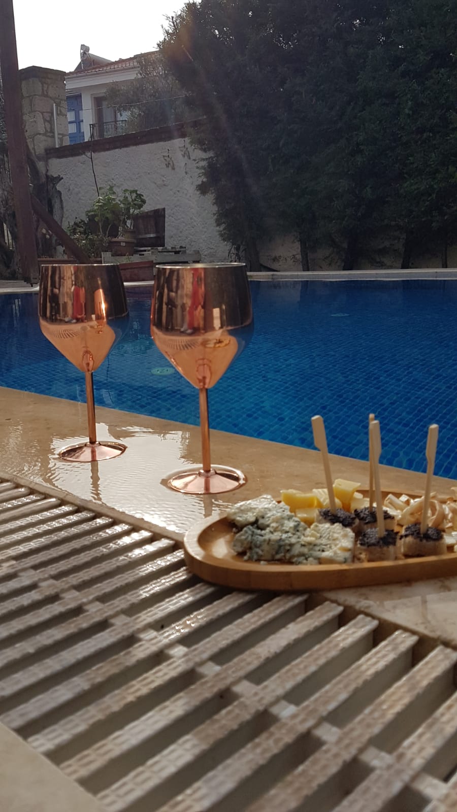 Two elegant glasses with rose gold rims stand beside a wooden platter filled with assorted snacks. The platter is positioned on a wet, textured surface near a clear blue pool, reflecting the surrounding greenery and tranquil environment.