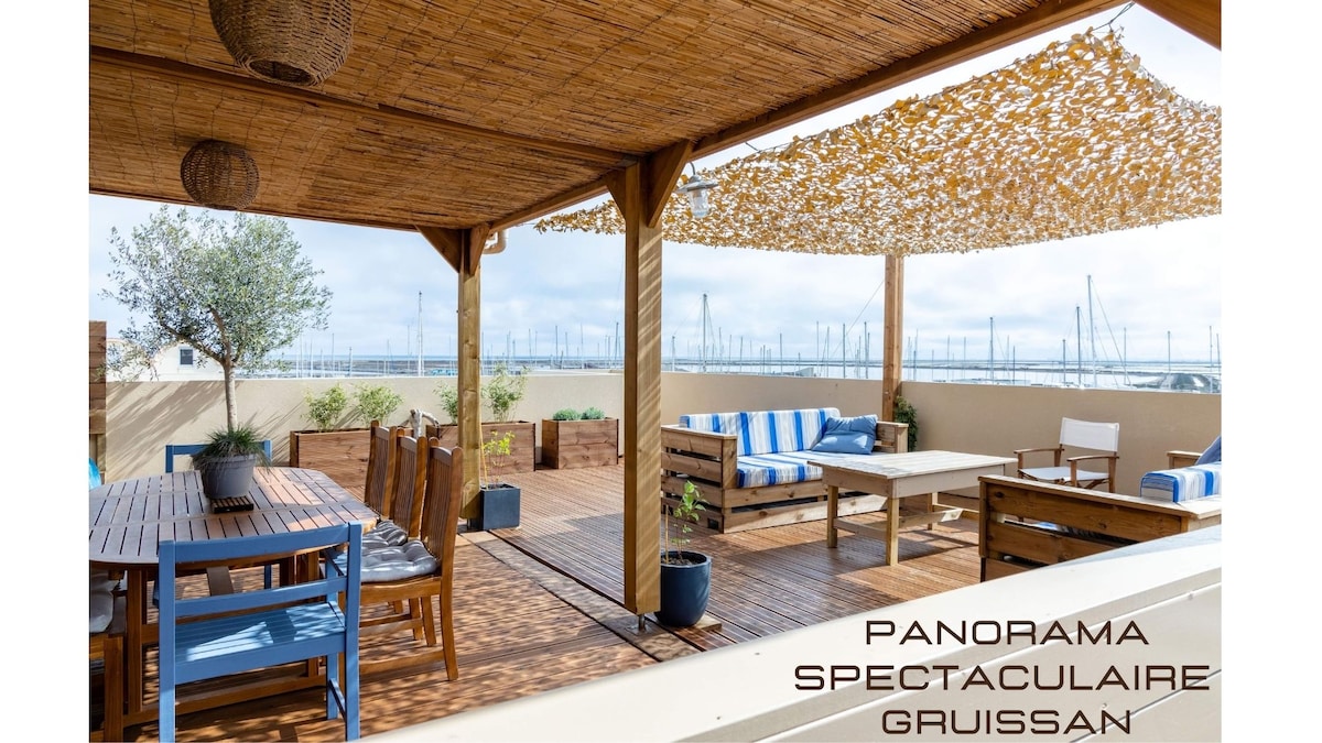 A spacious rooftop terrace features wooden decking, shaded lounge areas, and a dining table surrounded by comfortable seating. The area is adorned with natural elements, offering a panoramic view of the harbor, boats, and surrounding landscapes.
