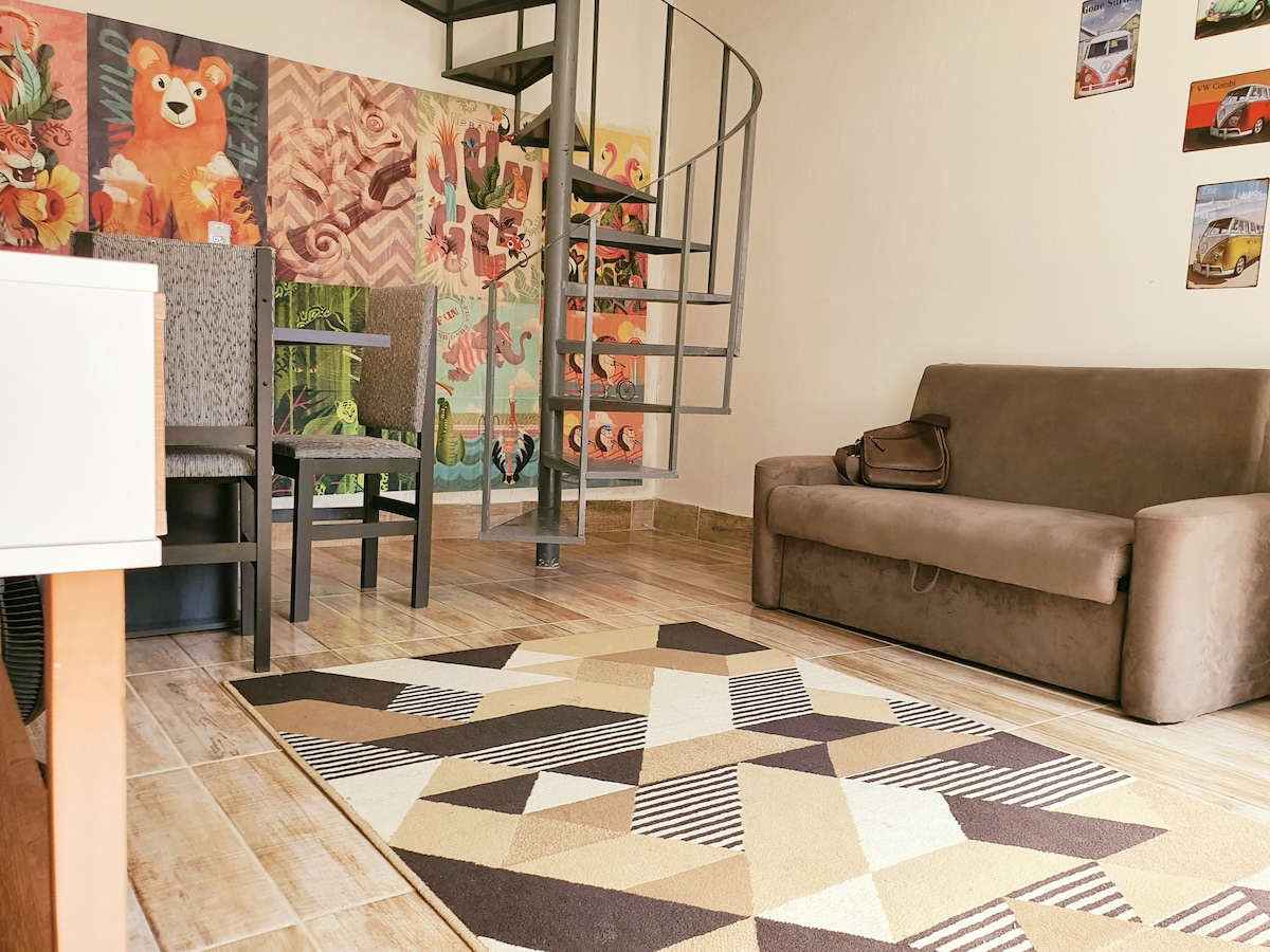 A living area features a spiral staircase leading to an upper level, with patterned artwork displayed on the wall. Two upright chairs and a couch in neutral tones complement a geometric area rug. Natural light brightens the space, creating a cozy atmosphere.