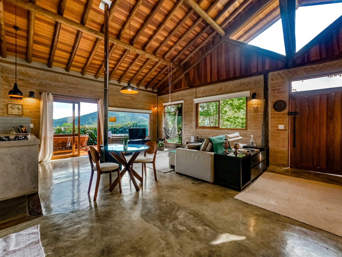 An open-plan living area features high wooden ceilings, large windows, and natural light. A comfortable sofa and a dining table are arranged. Views of the mountains and greenery are visible through the windows, enhancing the connection to nature.