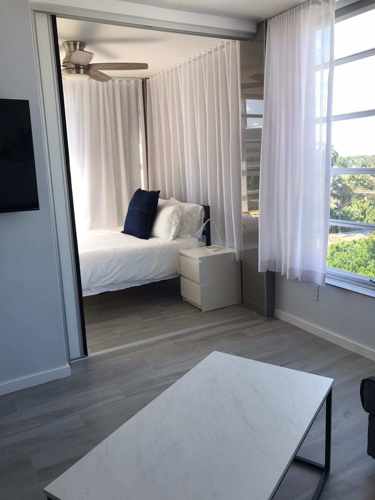 A cozy sleeping area featuring a queen bed dressed in white linens with a blue accent pillow. Light curtains frame the space, allowing natural light to filter through. A modern nightstand sits beside the bed, complementing the clean lines of the room.