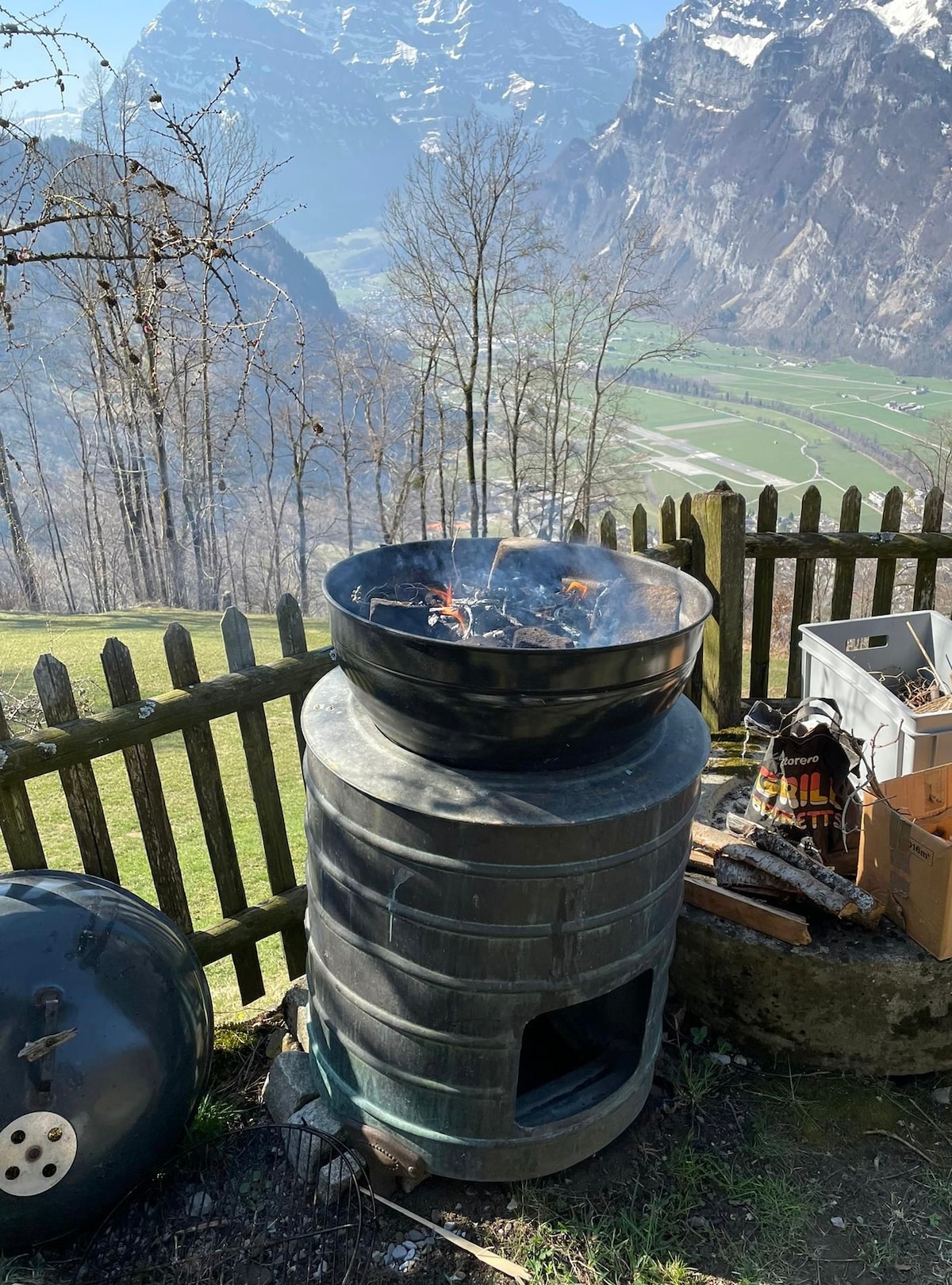 A charcoal grill stands on a stone base, with flames visible inside. The surrounding area is open, offering expansive views of the mountains and valleys in the background. A wooden fence encloses the space, enhancing the natural setting.