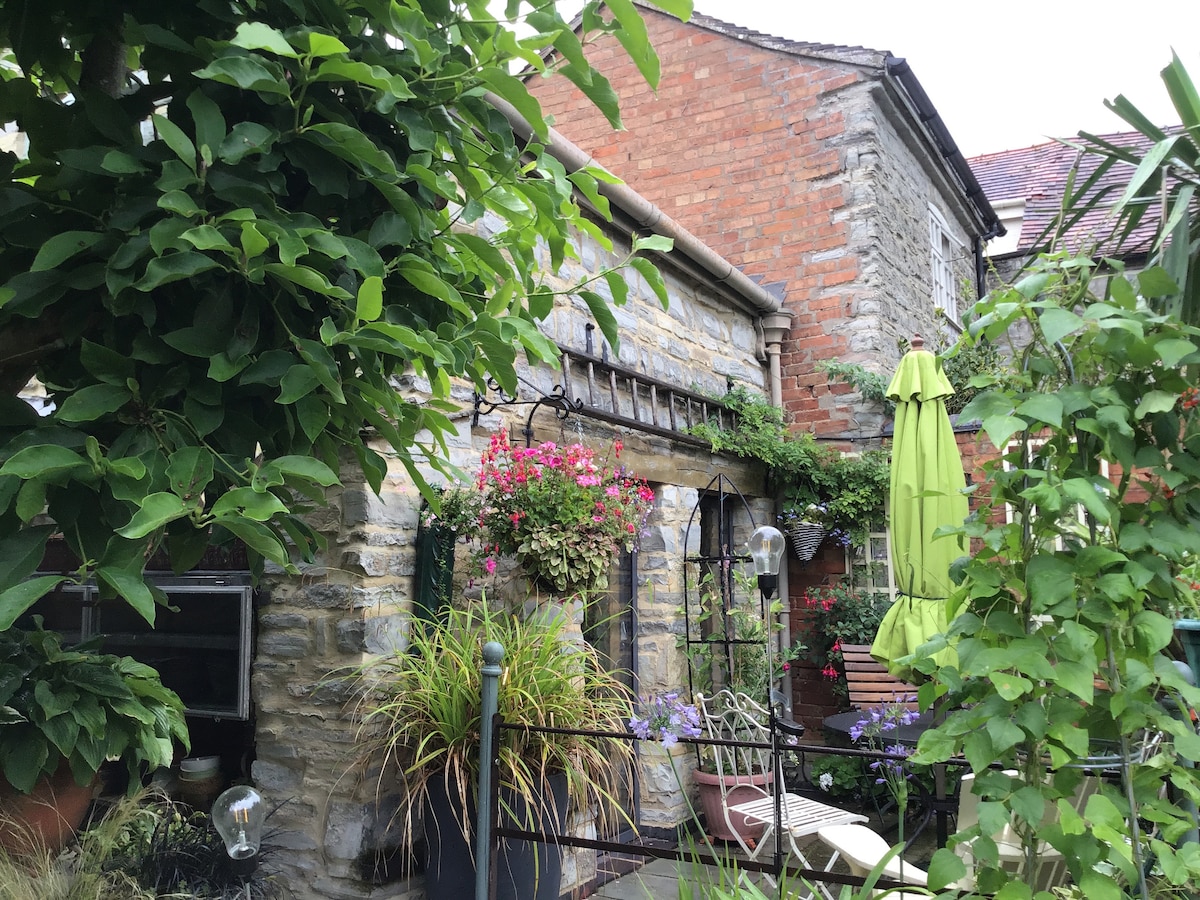 A charming stone exterior is framed by lush greenery and vibrant flowers. Potted plants and hanging blooms add color to the space, while a green umbrella provides shade over a seating area. The rustic details enhance the welcoming atmosphere of the outdoor courtyard.