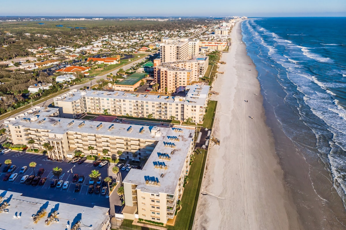 An aerial view showcases the beachfront property, featuring multiple buildings along the coastline. Sandy beaches extend towards the horizon, with gentle waves lapping at the shore. The area is bordered by greenery and a road, with various vehicles parked in the lot.