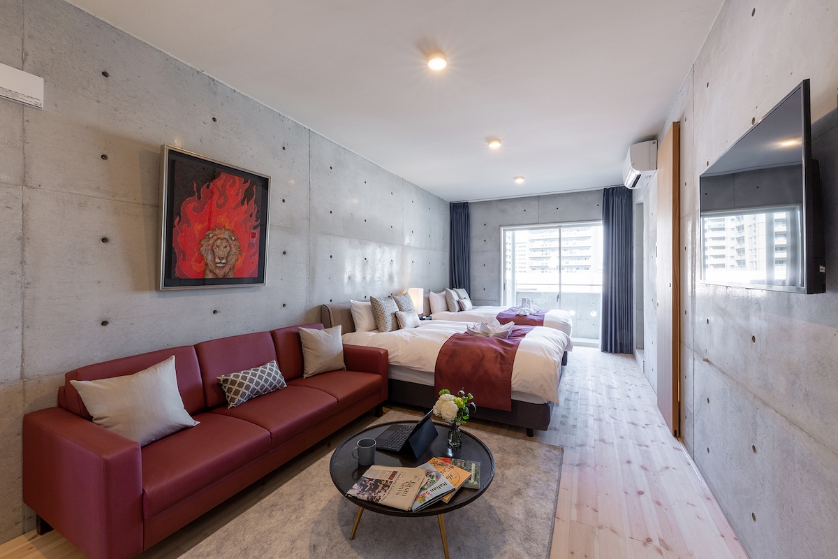 A spacious room features a comfortable seating area with a red sofa and a coffee table. Two expansive beds are positioned against a concrete wall, accompanied by decorative pillows. Natural light enters through a large window, and a colorful art piece highlights the wall.