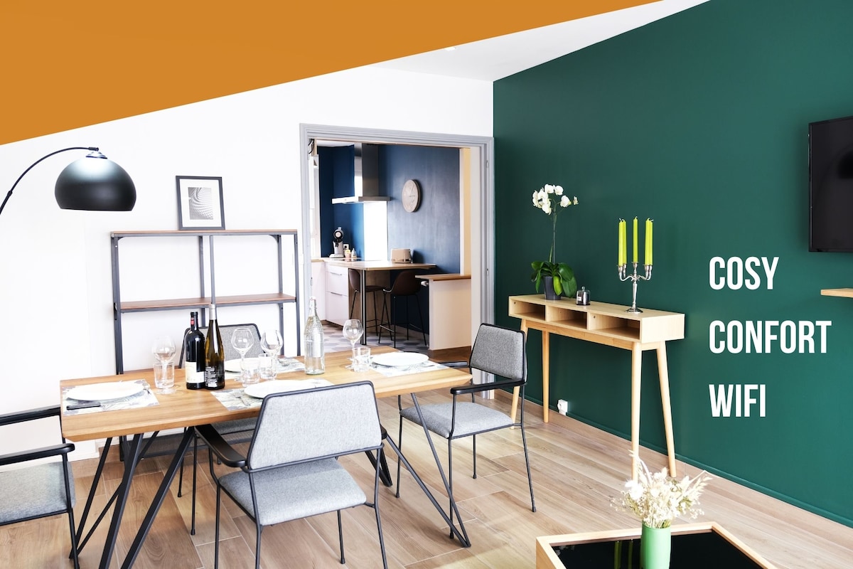 A modern dining area is showcased, featuring a wooden table set with dishes and glassware. Grey chairs are arranged around the table. A green accent wall adds a pop of color. A wall-mounted TV is visible adjacent to a console table.