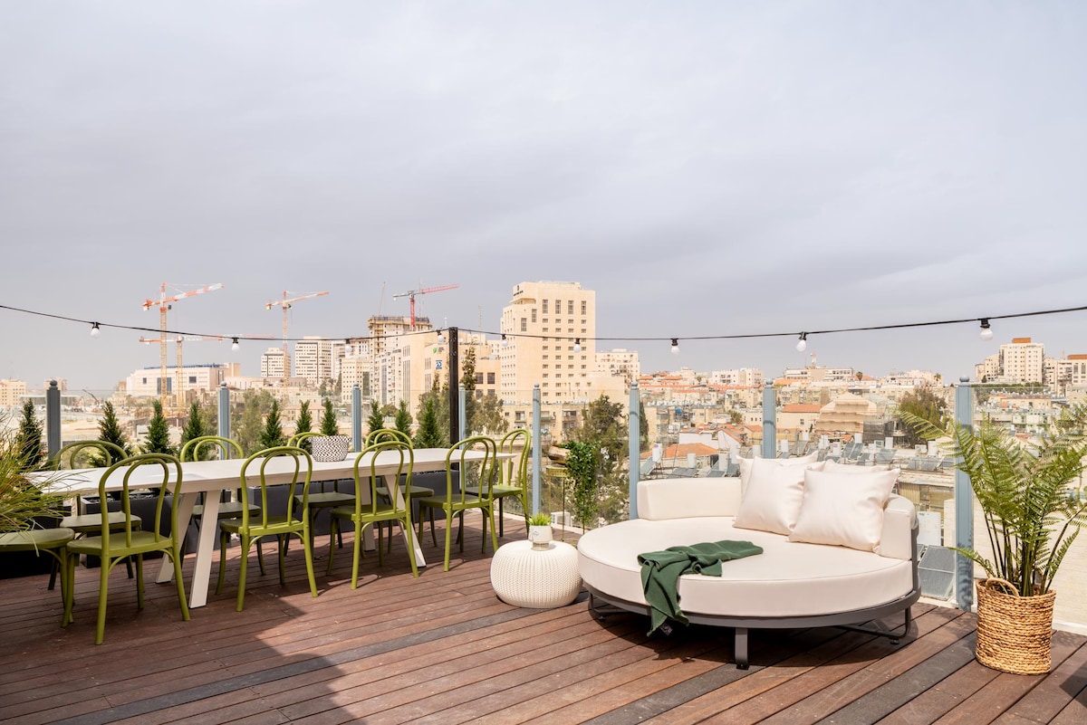 A spacious outdoor terrace is shown, featuring a circular seating area and a long dining table surrounded by green chairs. Potted plants are arranged along the perimeter, with cityscape views visible in the background under a cloudy sky.