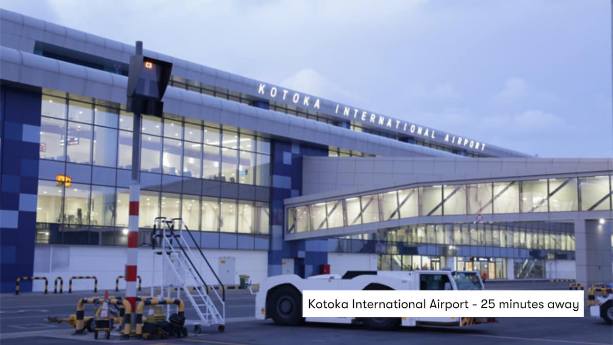 Kotoka International Airport is visible, featuring a modern architectural design with large glass windows. The exterior reflects soft evening light, while airport vehicles and equipment are stationed nearby. The location is noted to be approximately 25 minutes from the apartment.