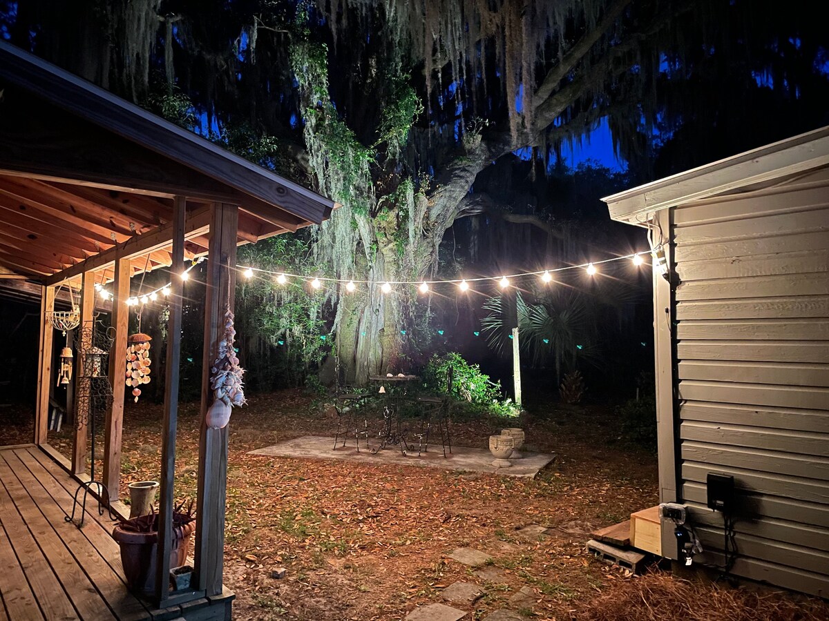 A serene outdoor space is illuminated by string lights, casting a warm glow against the backdrop of a large moss-draped tree. The area is surrounded by nature, with a stone patio and a dining set visible, creating an inviting atmosphere for gatherings.