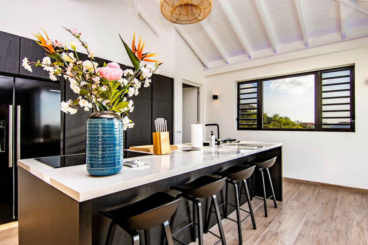 A modern kitchen features a large island with seating for four, complemented by a crisp white countertop. A blue vase filled with vibrant flowers adds a pop of color. Bright natural light fills the space through a large window with wooden slats.