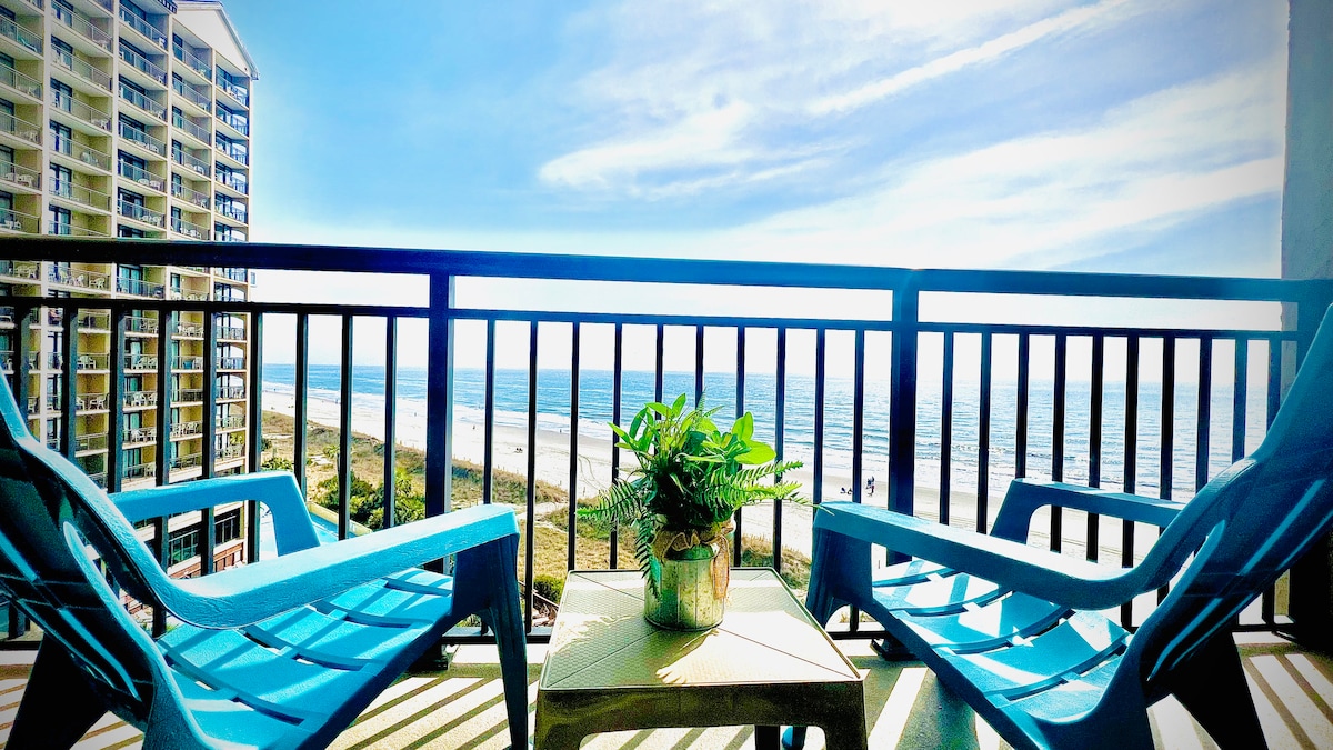 A private balcony features two blue chairs and a small table, with a potted plant at its center. The ocean and sandy beach stretch out into the distance, framed by clear skies and gentle waves, providing a serene coastal view.