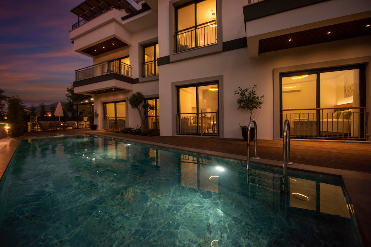 The exterior view highlights a modern building featuring large glass windows and balconies overlooking a swimming pool. Soft lighting illuminates the pool area, creating a serene ambiance. Potted plants are positioned near the pool, enhancing the outdoor space.