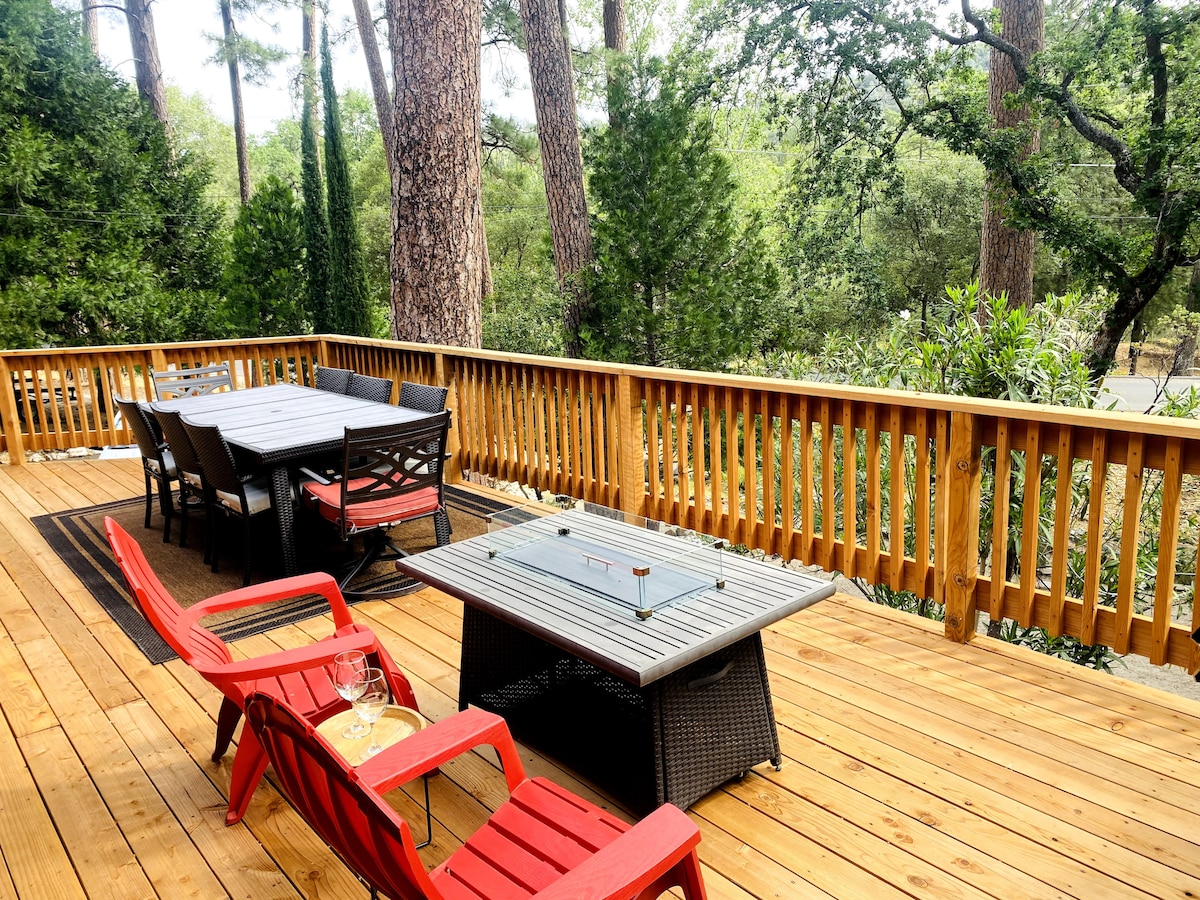 A spacious deck with wooden flooring is surrounded by tall trees, offering a serene outdoor space. A large dining table accommodates several guests, with additional seating provided by red chairs. A stylish coffee table adds a functional centerpiece to the gathering area.