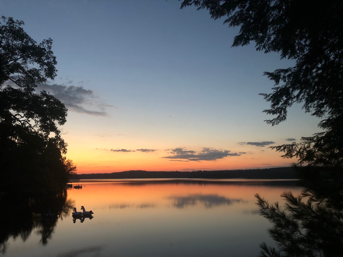 A serene sunset over Lake St. George reflects vibrant colors on the water's surface. Silhouettes of trees frame the view, while a few boats are spotted in the distance, contributing to the tranquil landscape.