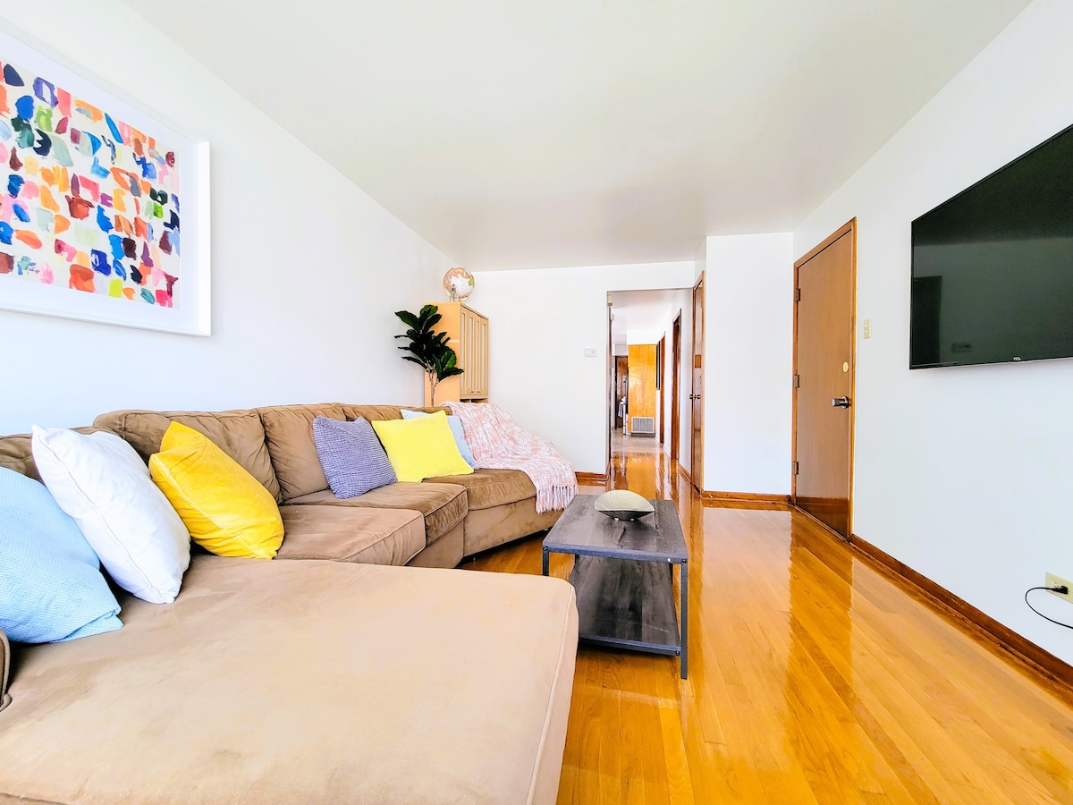 A comfortable living room is showcased, featuring a sectional sofa adorned with colorful cushions. A wooden coffee table sits in front of the sofa, while a flat-screen TV is mounted on the wall. Natural light fills the space, accentuating the warm wooden floors.