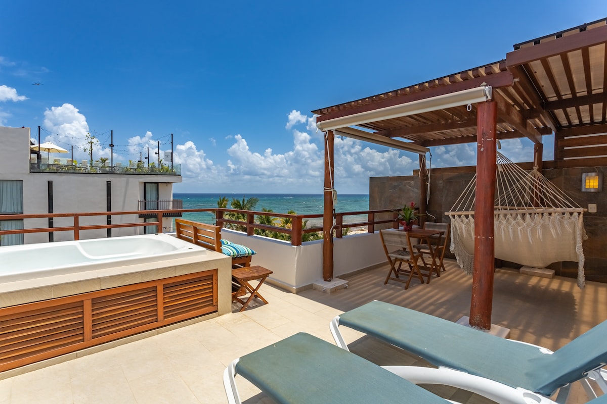 A spacious private roof terrace features a Jacuzzi with a view of the ocean. Two sun loungers are positioned nearby, complemented by a wooden table and folding chairs. A hammock is also present, creating a relaxing outdoor space surrounded by tropical greenery.