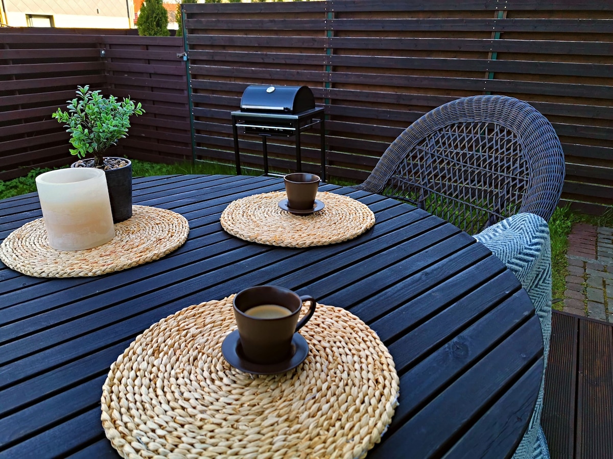 A circular outdoor table is set with woven placemats and dark cups, accompanied by a neutral-toned candle. A potted plant and a small grill are positioned in the background, while textured chairs provide comfortable seating. The enclosed space offers privacy and a relaxed atmosphere.