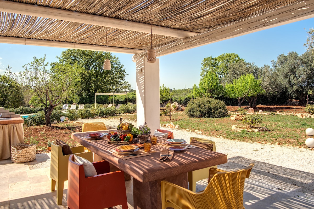 An outdoor dining area is shaded by a wooden pergola, featuring a substantial table set with plates and colorful decorative fruits. Surrounding the table are comfortable chairs, with a scenic view of a lush garden that includes trees and flowering plants.