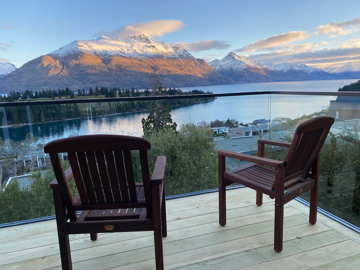 Two wooden chairs are positioned on a balcony, overlooking a serene lake and majestic mountains. The scene features subtle morning light casting a gentle glow on the landscape, highlighting the snow-capped peaks and tranquil waters.