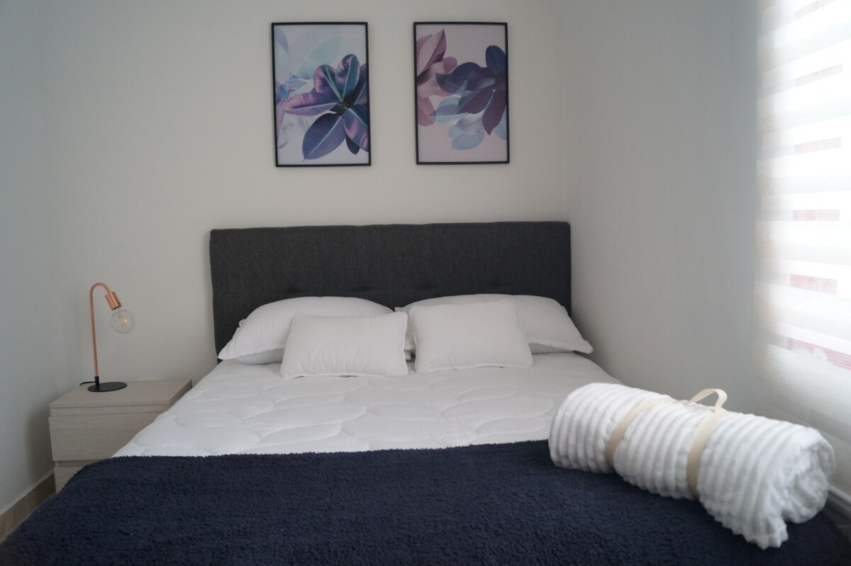 A sleeping area features a spacious bed with white linens and two decorative pillows. Above the bed, two framed floral artworks are displayed. A bedside table with a modern lamp is positioned on one side, while a neatly rolled towel rests on the opposite side.