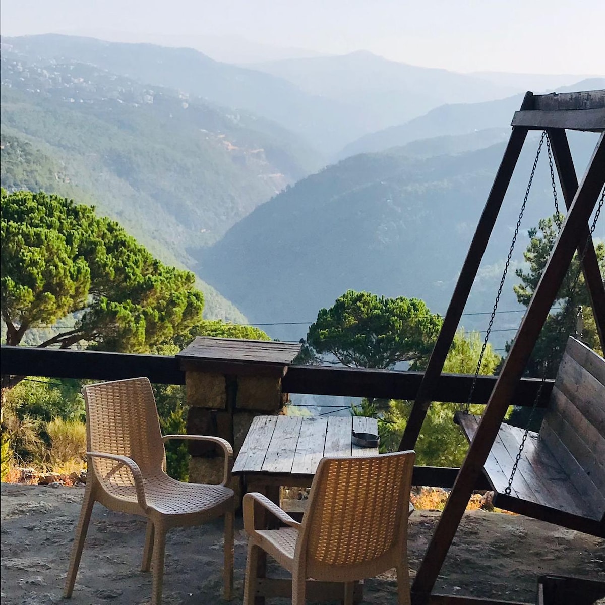 A serene outdoor seating area features two chairs and a wooden table, with a swing set nearby. The view overlooks a mountainous landscape, showing layers of green hills under a hazy sky.