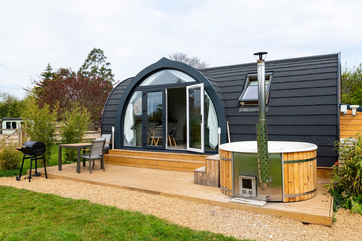 An exterior view of a stylish, modern accommodation showcases large glass doors leading to a spacious deck. A wood-fired hot tub is situated next to a wooden table and chairs. Green grass and landscaped foliage create a serene outdoor setting.