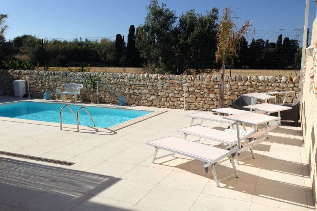 A swimming pool is framed by a spacious tiled patio featuring several lounge chairs. A stone wall borders the area, and greenery is visible in the background, creating a serene outdoor setting. Sunlight enhances the inviting ambiance around the pool.