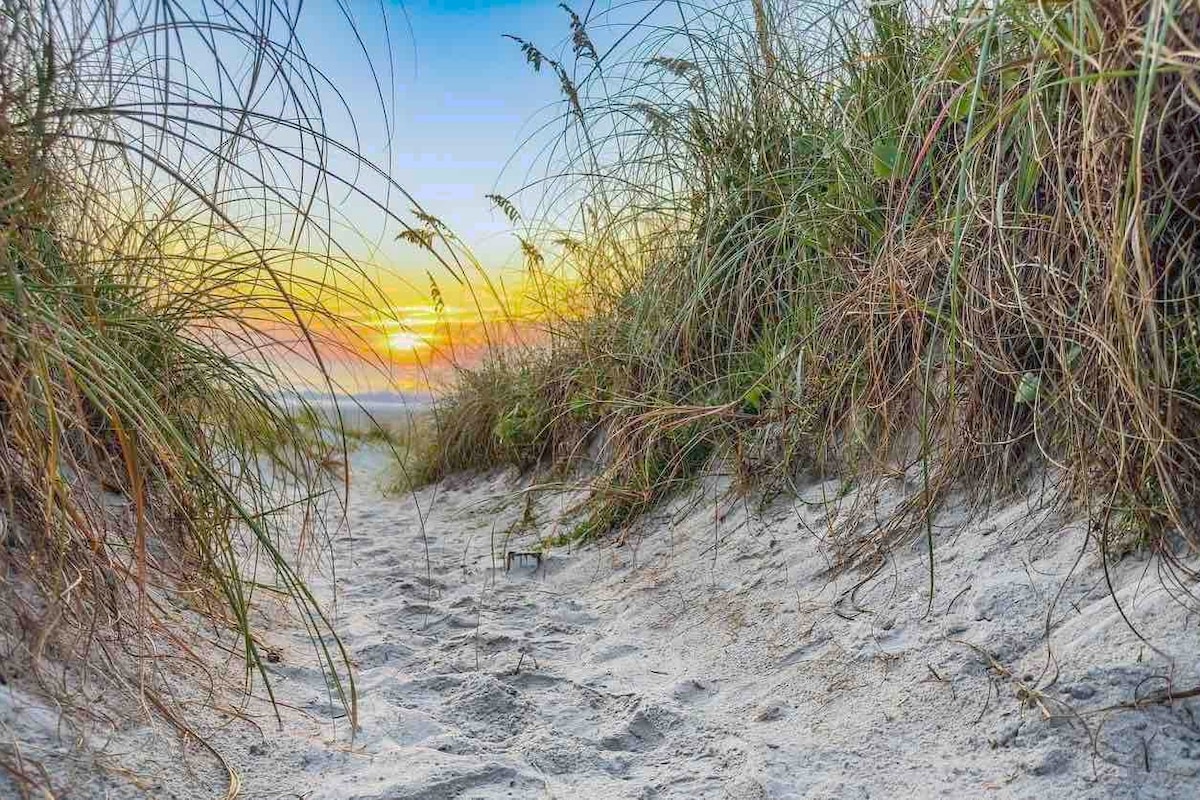 A sandy pathway through tall grass leads to the beach, where the sun is rising above the horizon, casting warm colors across the sky. Gentle grasses sway in the foreground, framing the serene coastal view.