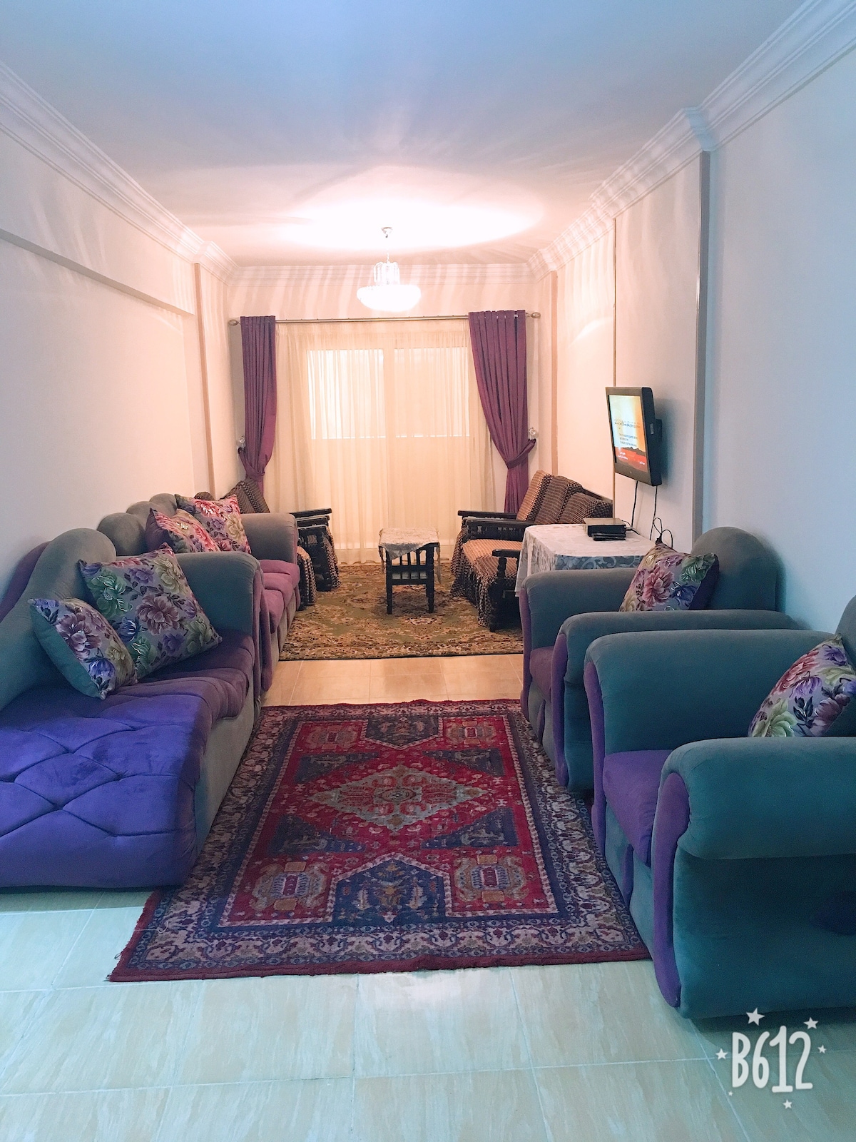 A shared living space is presented, featuring several plush chairs and a cozy sofa, arranged around a central area rug. Natural light floods in through large windows, with decorative curtains framing the view. A small dining table is located at the far end of the room.