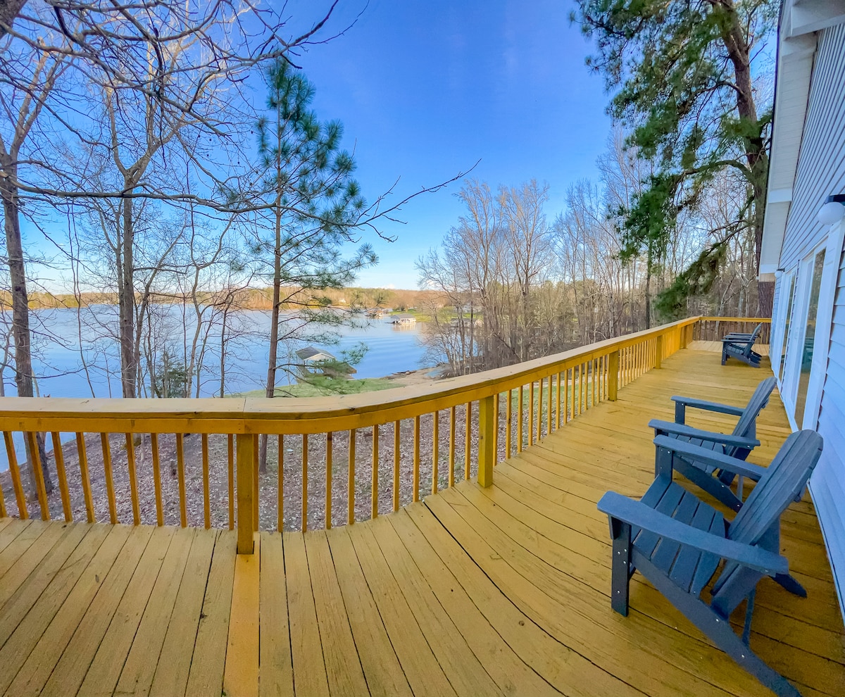 A spacious wooden deck is lined with Adirondack chairs, offering views of the lake and surrounding trees. The setting captures a tranquil atmosphere, with the water reflecting the clear sky, creating a serene backdrop for relaxation.