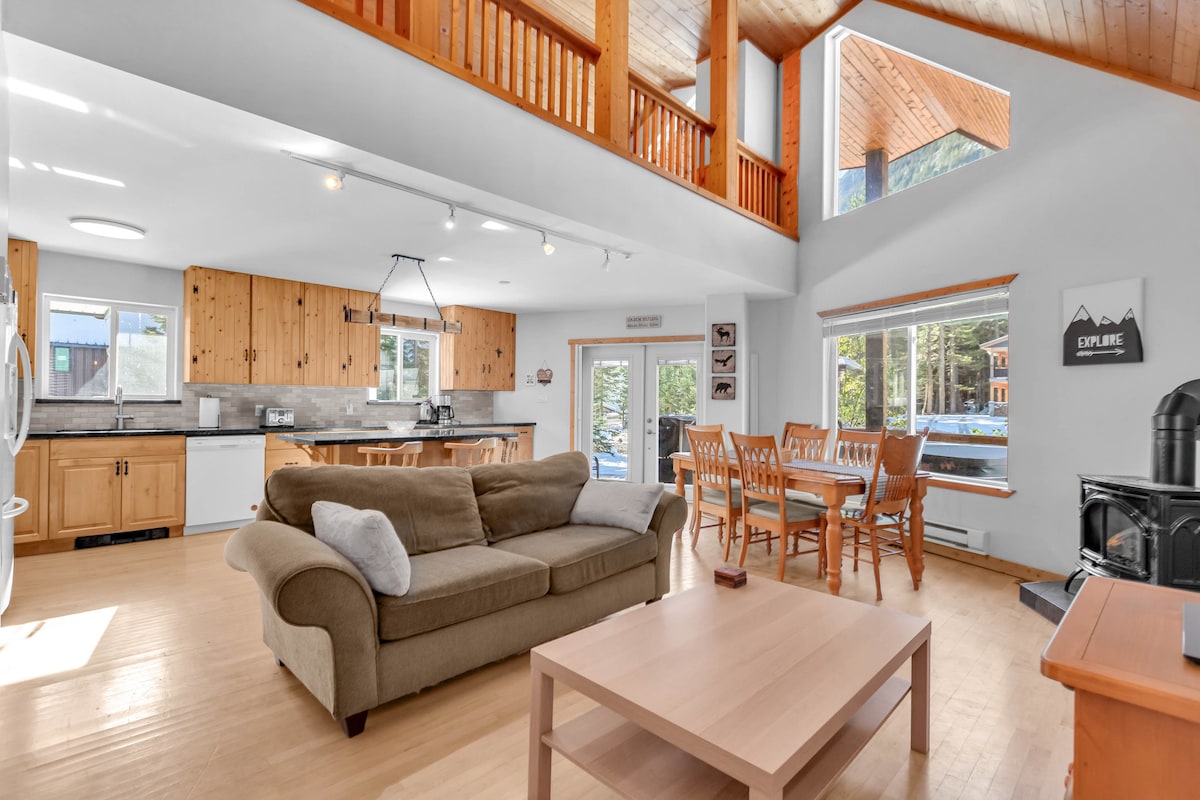 An open-concept living area features natural wooden accents and spacious windows allowing for ample natural light. A comfortable couch is positioned near a coffee table, with a dining table and chairs visible in the background, creating an inviting space for relaxation and gatherings.