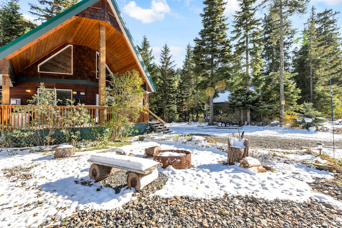 A rustic chalet is surrounded by a tranquil outdoor setting covered with a light layer of snow. Stone pathways lead to a wooden bench and a firepit, nestled among scattered trees, inviting relaxation in nature.