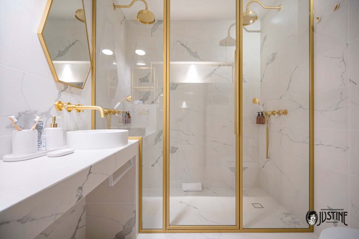 A luxurious bathroom features a spacious double shower enclosed in glass. Elegant gold fixtures complement the stylish marble tiles. A well-lit mirror above a modern sink adds functionality, while bottles of toiletries are neatly displayed on the adjacent shelf.