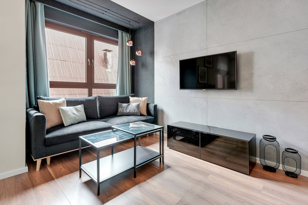 A modern living area features a dark sofa accented with light cushions, positioned near a large window that brings in natural light. A flat-screen TV is mounted on the wall opposite the sofa, and a coffee table set is placed in front of it, adding to the room's cozy atmosphere.