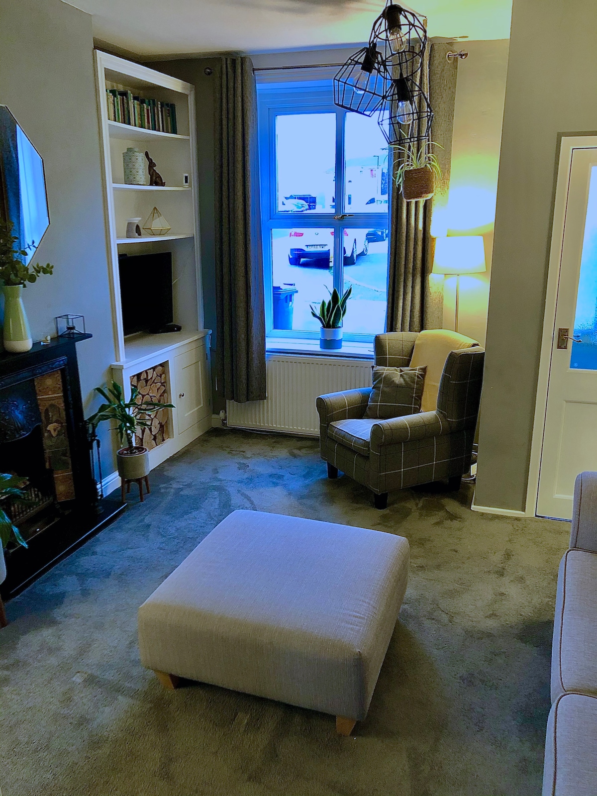 A cozy living room features a comfortable armchair beside a window, allowing natural light to fill the space. A small ottoman rests on the carpeted floor, while a fireplace is adorned with logs and plants. Bookshelves line one wall, adding a homely touch.