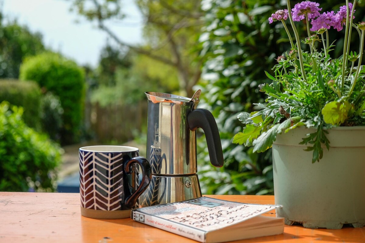 A sunlit outdoor setting features a shiny coffee maker alongside a patterned mug resting on a wooden table. A light-colored book lies open next to the cup, while a potted plant with purple flowers adds a touch of color in the background.